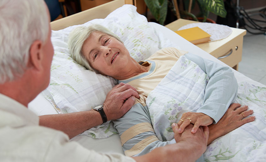 An older man comforts an older woman who is lying in bed looking at him
