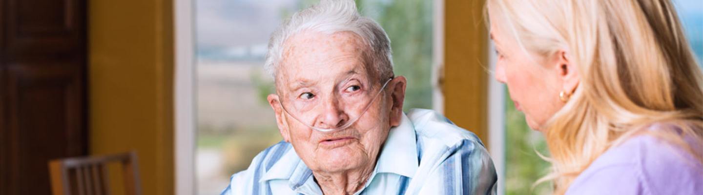 An older man with a nasal cannula for oxygen talks with his daughter at home