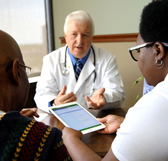 A physician seated at a table talks with two people while they look at a tablet