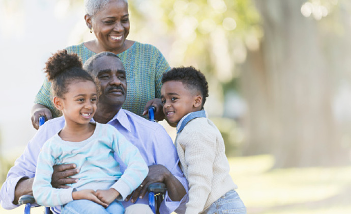 A man seated in a wheelchair has his granddaughter in his lap while surrounded by his wife and grandson