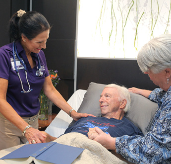 A man lying in bed and his wife, standing next to him, talk with a VITAS team member