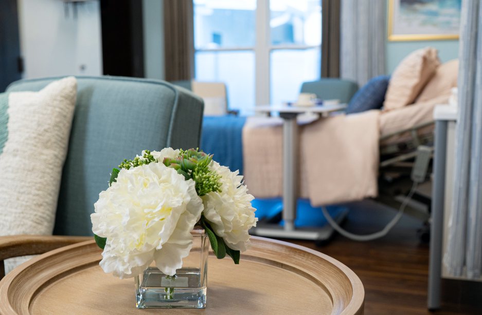 A small vase of flowers on a table in the foreground of a sunny room with a hospital bed with pillows and blankets in the background